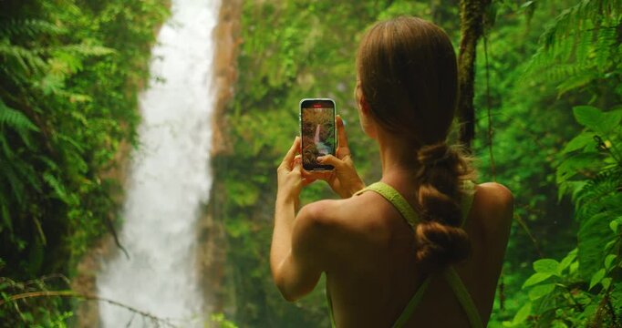 Tourist filming tall Costa Rica waterfall surrounded by lush rainforest and Blue Morpho butterfly flying near her