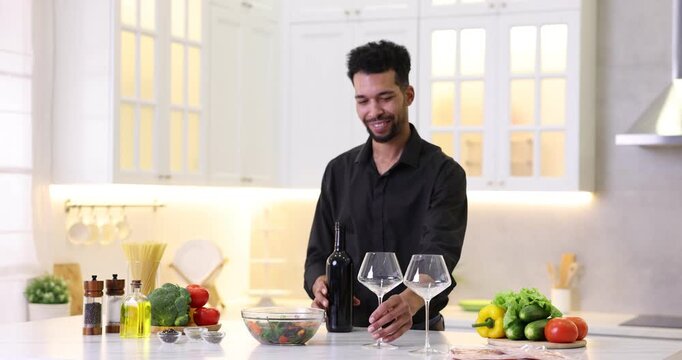 Man opening wine bottle with corkscrew and pouring drink into glass at table in kitchen