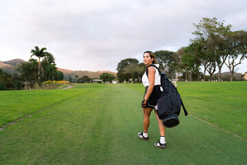Latin hispanic woman smiles looking back while carrying golf bag on golf course