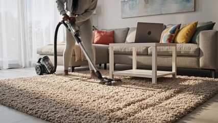 A person vacuums a sunlit living room with a canister cleaner as sofa and laptop sit nearby in view!