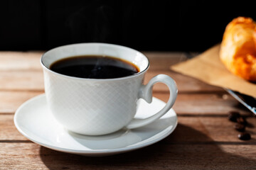 Black coffee in a white cup, roasted coffee beans and toast placed on a table in the morning sunlight, against a backdrop of dark black wooden walls.
