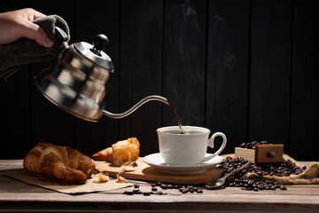 Coffee is poured into a white coffee cup. Roasted coffee beans and toast are placed on the table in the morning sunlight, against a backdrop of dark wooden walls.