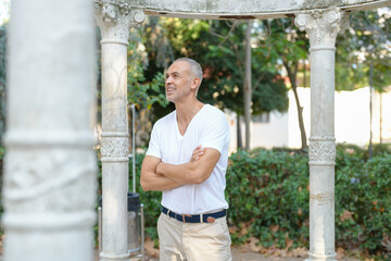 A man in a white shirt and khaki pants stands in front of a stone archway. He is smiling and he is...