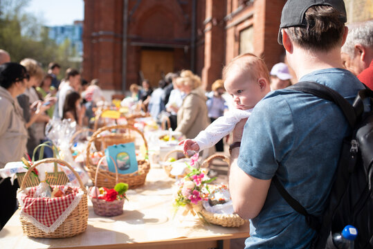 A baby in his father's arms. A father and son stand in front of a table with many baskets of Easter food for blessing next to the church.