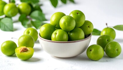 Bowl of unripe green plums, some scattered, with leaves on a white surface, one halved, revealing a light pit