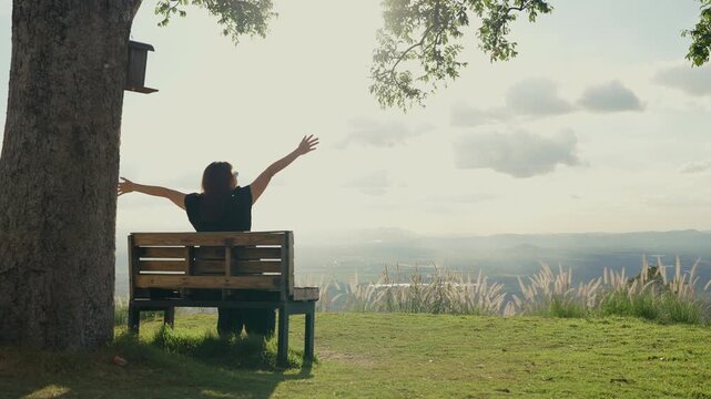Rear view of a woman sitting alone on a wooden bench under a large tree, enjoying the mountain view, breathing in fresh air, and immersed in a peaceful natural atmosphere