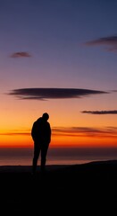 Fototapeta premium Solitary figure stands silhouetted against a vivid twilight horizon over water
