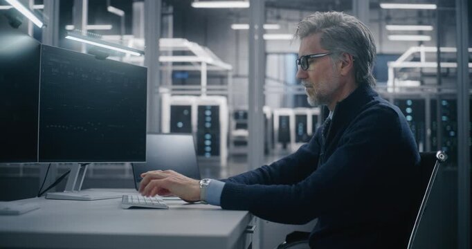 Focused, Middle Aged Male Senior Engineer Typing on Keyboard, Monitoring Cloud System Metrics on Dual Screens in Data Center. Critical Infrastructure Analysis and Cloud Infrastructure Management.