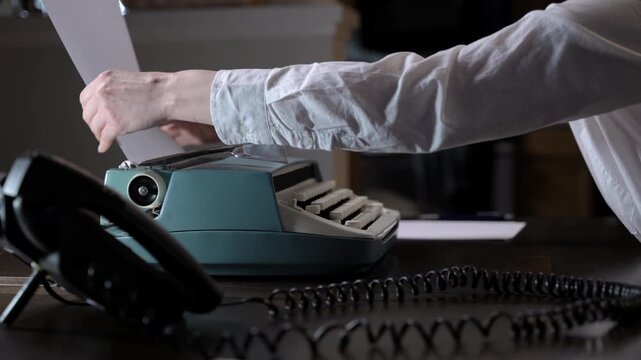 Classic typewriter on a desk with a paper inserted, next to a retro telephone. Hands working in the background, creating a nostalgic office and writing atmosphere.