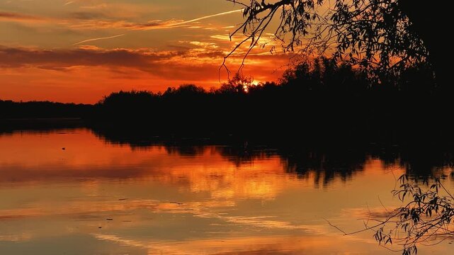 A colorful sunset in the picturesque sky over the Volga near Astrakhan. The sun disk enters the water. A beautiful summer or spring sunset on the Volga with trees reflected in the water. 4К