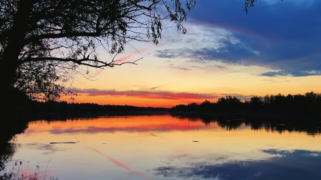 A colorful sunset in the picturesque sky over the Volga near Astrakhan. The sun disk enters the water. A beautiful summer or spring sunset on the Volga with trees reflected in the water. 4К