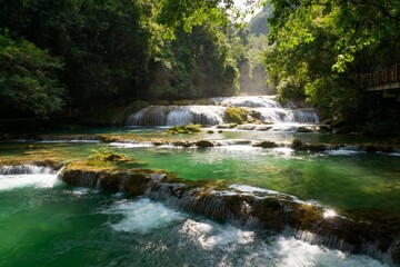 A serene cascade of waterfalls flowing through a lush green forest with clear turquoise water and moss-covered rocks.