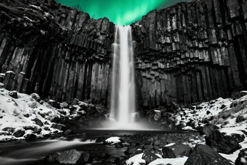 A cascading waterfall framed by basalt columns under a green aurora in a snowy landscape