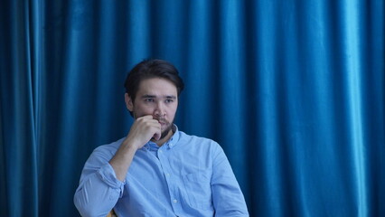 Pensive young man in deep contemplation looking away, sitting against a blue background.