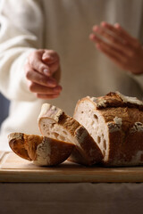 A rustic loaf of sourdough bread is placed on a wooden cutting board, with a woman wearing a soft white sweater in the background