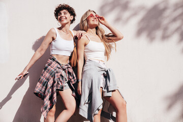 Two young beautiful smiling hipster female in trendy summer white t-shirt and shorts clothes. Sexy carefree women posing in the street. Positive models having fun, hugging and going crazy, near wall
