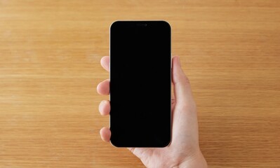 Hand holding a smartphone with blank screen on a wooden table viewed from above