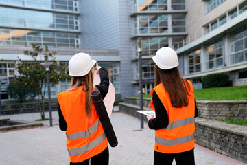 Two women engineers wearing hard hats and safety vests reviewing blueprints and plans at a modern...
