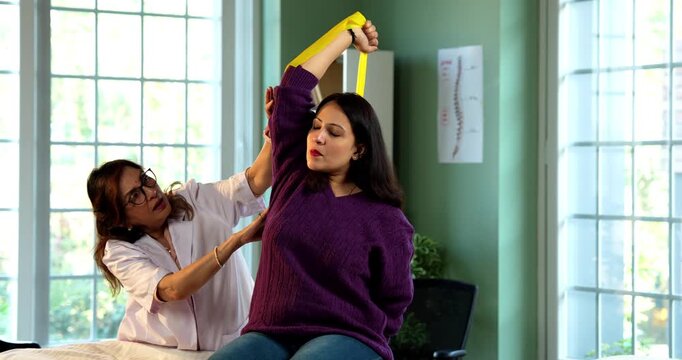 Indian lady patient with physiotherapist performing triceps and shoulder strengthening exercises using resistance band in modern clinic under senior guidance for rehabilitation and recovery