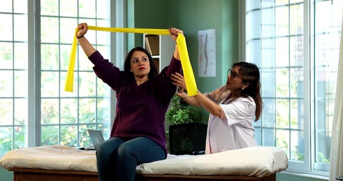 Indian lady patient with physiotherapist performing triceps and shoulder strengthening exercises using resistance band in modern clinic under senior guidance for rehabilitation and recovery