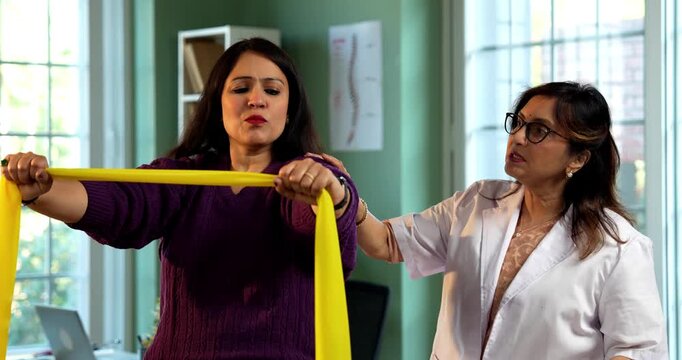 Indian lady patient with physiotherapist performing triceps and shoulder strengthening exercises using resistance band in modern clinic under senior guidance for rehabilitation and recovery