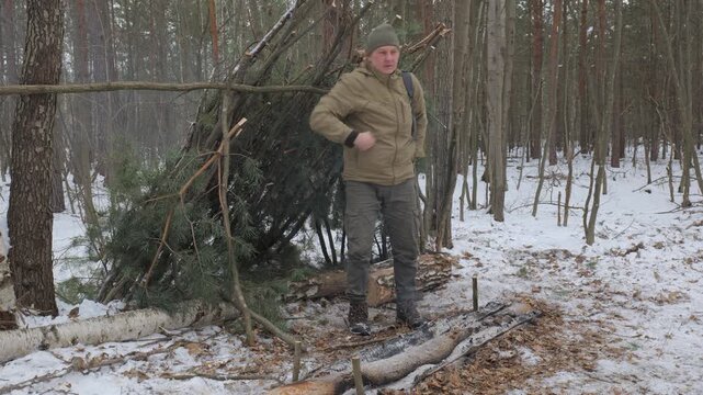 A bushcraft tourist near a primitive shelter he built in a snowy winter forest, a shelter made of branches, designed for survival during hikes in the cold season.