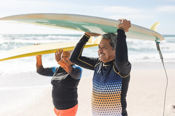 Naklejka premium Happy multiracial senior couple carrying surfboards over heads at beach during sunny day