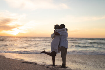 Side view of senior multiracial couple embracing and kissing on shore at beach during sunset