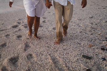 Naklejka premium Low section of senior multiracial couple holding hands walking barefoot on sand at beach