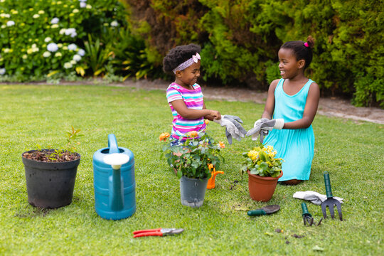 Playful smiling african american sisters kneeling while wearing gloves by plants in garden - Powered by Adobe