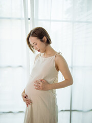 Happy Asian pregnant woman lovingly touching her unborn baby while standing by the window at home.