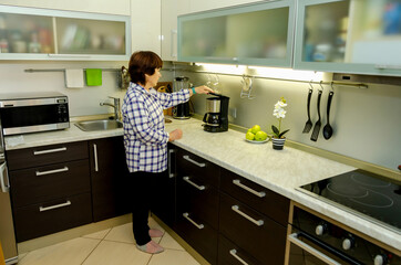 Elderly woman turns on the coffee maker to make coffee in modern kitchen, calm morning ritual, domestic independence, everyday comfort, quiet self-care, technology use.