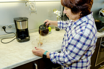 Elderly woman pouring fresh coffee from glass pot into clear mug in modern kitchen, calm morning routine, home comfort, cozy domestic lifestyle, warm lighting, everyday moments.