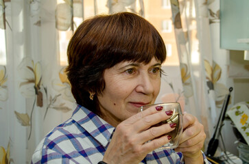 Elderly woman smiling gently while holding clear glass mug of coffee near her lips, inhaling the fragrance. soft natural light, domestic moment, contentment, home comfort, warm atmosphere.