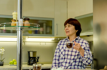Elderly woman sipping hot coffee from glass cup while standing at kitchen counter near coffee maker, relaxed pose capturing morning enjoyment amid appliances. Home leisure moment.