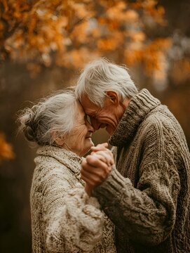 An elderly couple in warm sweaters dances together outdoors du autumn with colorful leaves falling from the trees above enjoying their day.