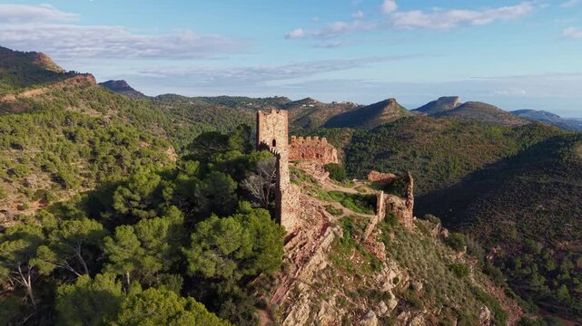 Cinematic aerial orbit reveals the remains of a historic mountain castle near Serra village, surrounded by rugged terrain and pine forests in the Sierra Calderona Natural Park, Valencia, Spain