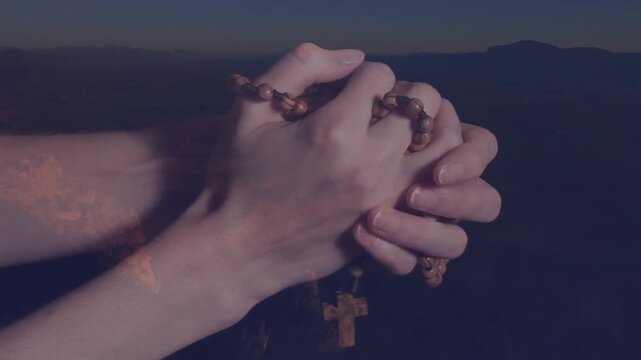 Clasping rosary beads, person praying over serene mountain landscape at dusk