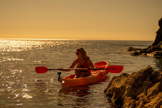 Woman, kayak, ocean. Woman paddles an orange kayak on sparkling water near rocky coastline at sunset with copy space. - Powered by Adobe