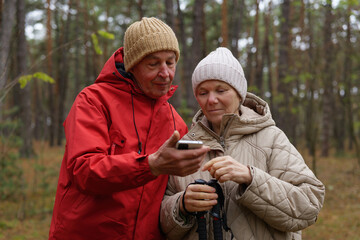 A couple stands together in a forest, checking their phone while dressed in warm jackets and hats. Surrounding trees show autumn colors under morning light