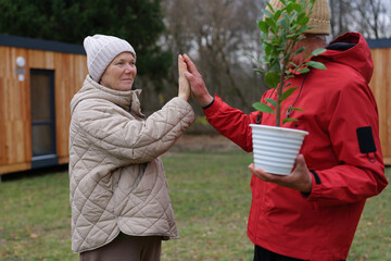Two elderly individuals greet each other in a garden. One person holds a plant in a pot. The trees...