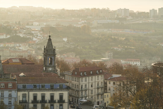 View of the cityscape with historic buildings, including a church steeple, under a soft, hazy sunlight glow, Porto, Porto District, Portugal.