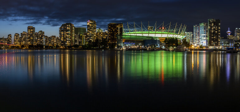 View of the vibrant city skyline with illuminated buildings and a green-lit stadium reflecting on the calm water at night, Vancouver, British Columbia, Canada.