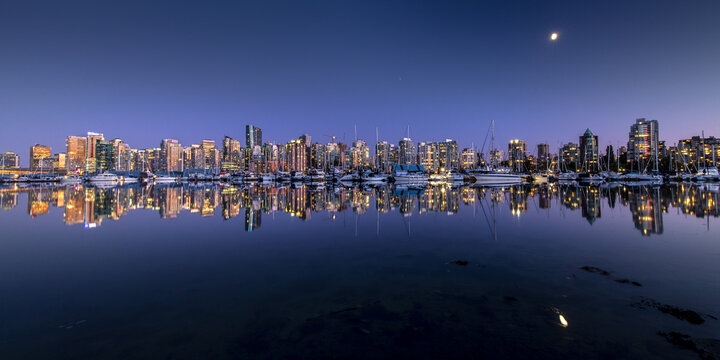 View of a city's skyline ablaze with golden lights, reflected in the still, dark water under a twilight sky with the moon above, Vancouver, British Columbia, Canada.