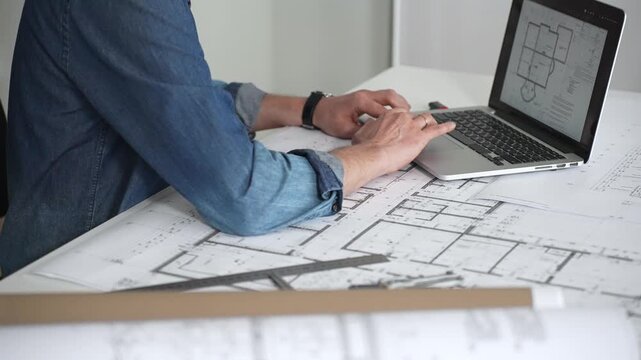 Architect sketching building plans on paper using a ruler and pen while working digitally on a laptop with a blueprint software displaying a floor plan