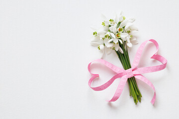 Naklejka premium Bouquet of snowdrop flowers with a bow of pink ribbon in the shape of hearts on a white background. Beautiful postcard, copy space