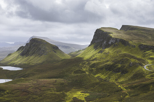 View of dramatic, rugged landscape with jagged peaks, emerald slopes, and winding roads under a moody, overcast sky, Isle of Skye, United Kingdom.