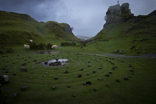 View of a stone circle nestles in a verdant valley under a moody sky, sheep graze peacefully nearby, and a solitary figure stands atop a rocky outcrop, Isle of Skye, United Kingdom.