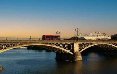 The red bus slowly crosses the Triana Bridge over the Guadalquivir River in Seville.