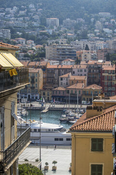 View of a vibrant cityscape unfolds with terracotta roofs cascading down to meet the turquoise harbor dotted with yachts, bathed in warm sunlight, Nice, France.